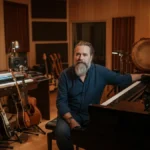 A male composer sits thoughtfully in his recording studio, surrounded by a piano, guitars, and world instruments.
