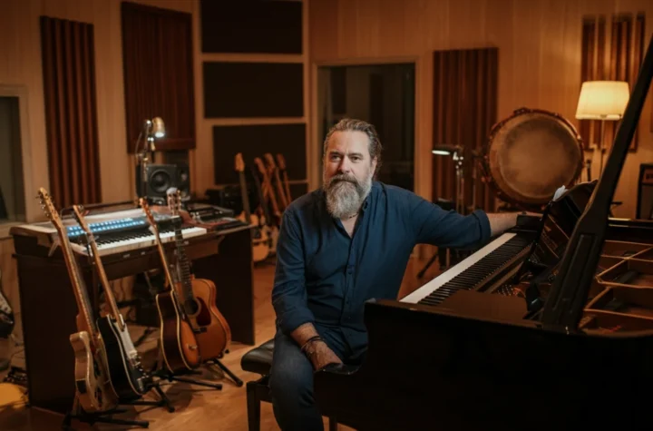 A male composer sits thoughtfully in his recording studio, surrounded by a piano, guitars, and world instruments.